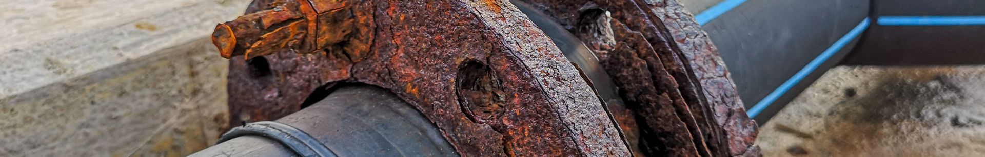 A close-up of a rusted metal flange connecting two black pipes, with a pier and water in the background